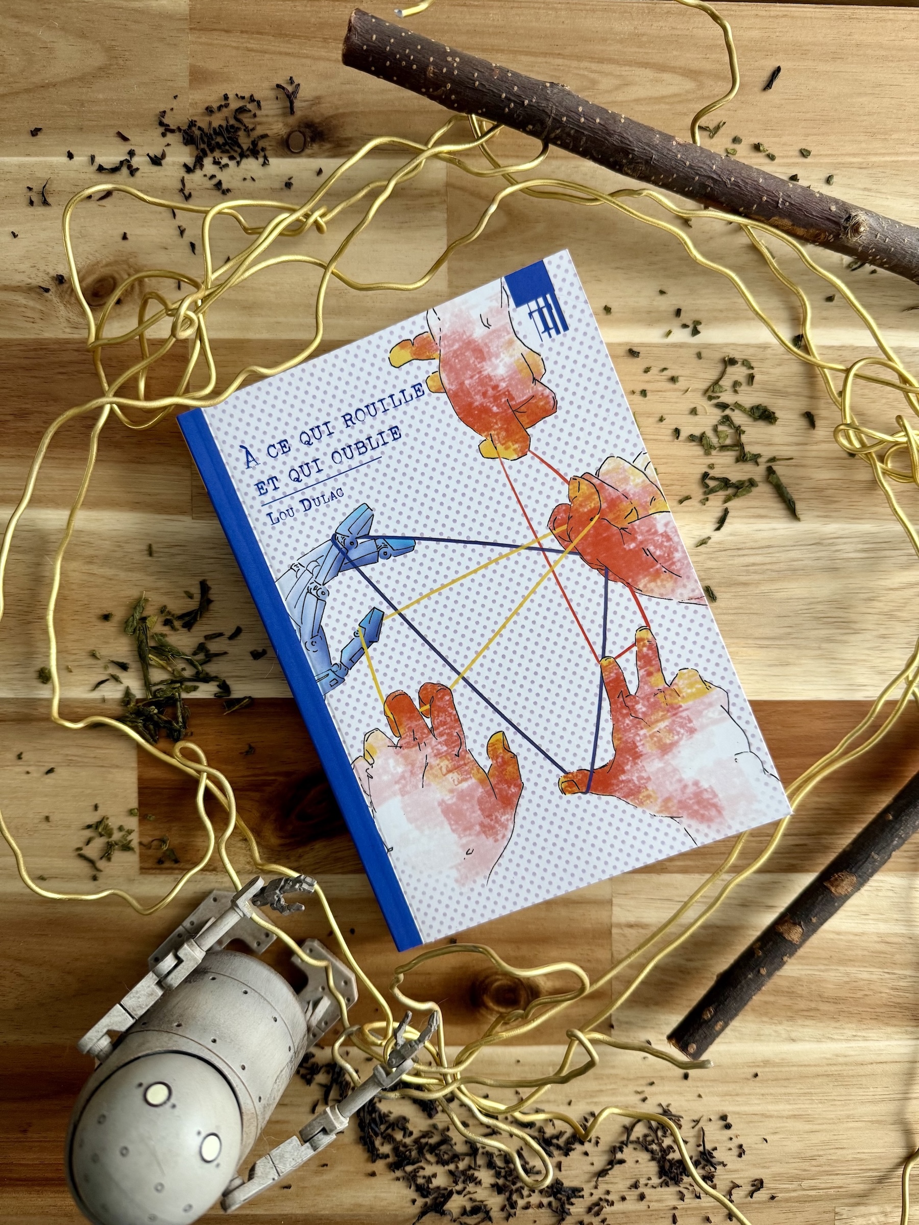 Livre posé entouré d'un fil de fer doré, de thé en vrac et avec des branches et un petit robot. La couverture de À ce qui rouille et qui oublie montre des mains humaines et une main robotique tenant entre eux des fils entre leurs doigts.