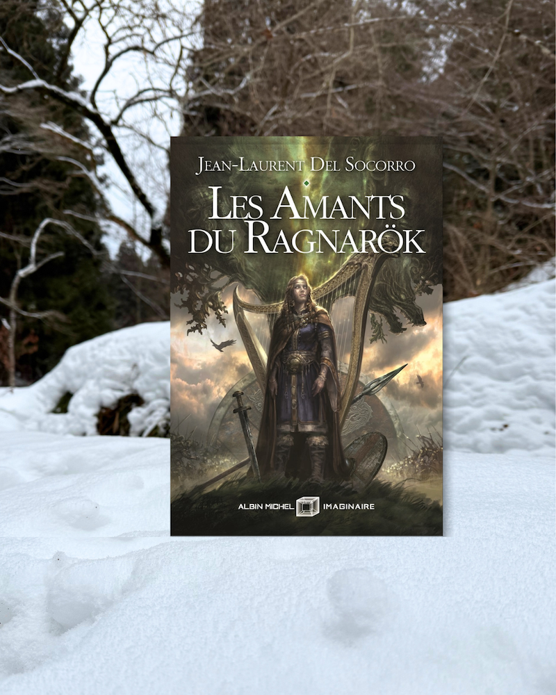 Livre pris en photo dans la neige dans un paysage forestier enneigé. La couverture des Amants du Ragnarök montre une scalde aveugle debout devant Yggdrasil, harpe dans le dos et lance fichée dans le sol devant elle avec épée et bouclier. En fond, deux armées qui fondent l'une sur l'autre