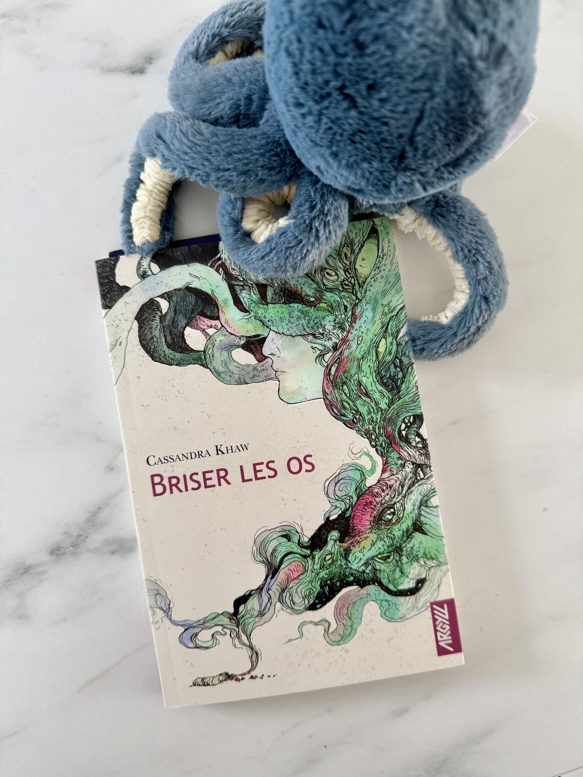 Livre posé à côté d'une peluche poulpe. La couverture montre un visage pris dans un mélange des tentacules et d'yeux.