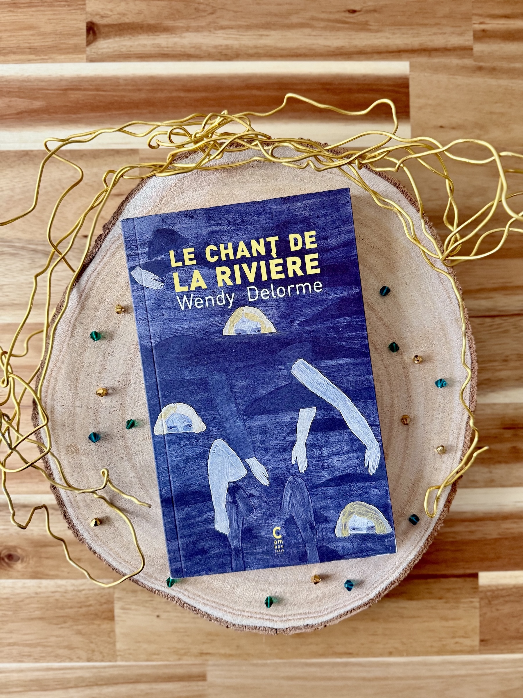 Livre posé sur un rondin de bois avec des perles de couleurs vertes et or et un fil doré torsadé doré. La couverture du livre montre, sur un fond bleu avec vagues des morceaux de corps et des visages aux cheveux blonds qui sortent à peine de l'eau.