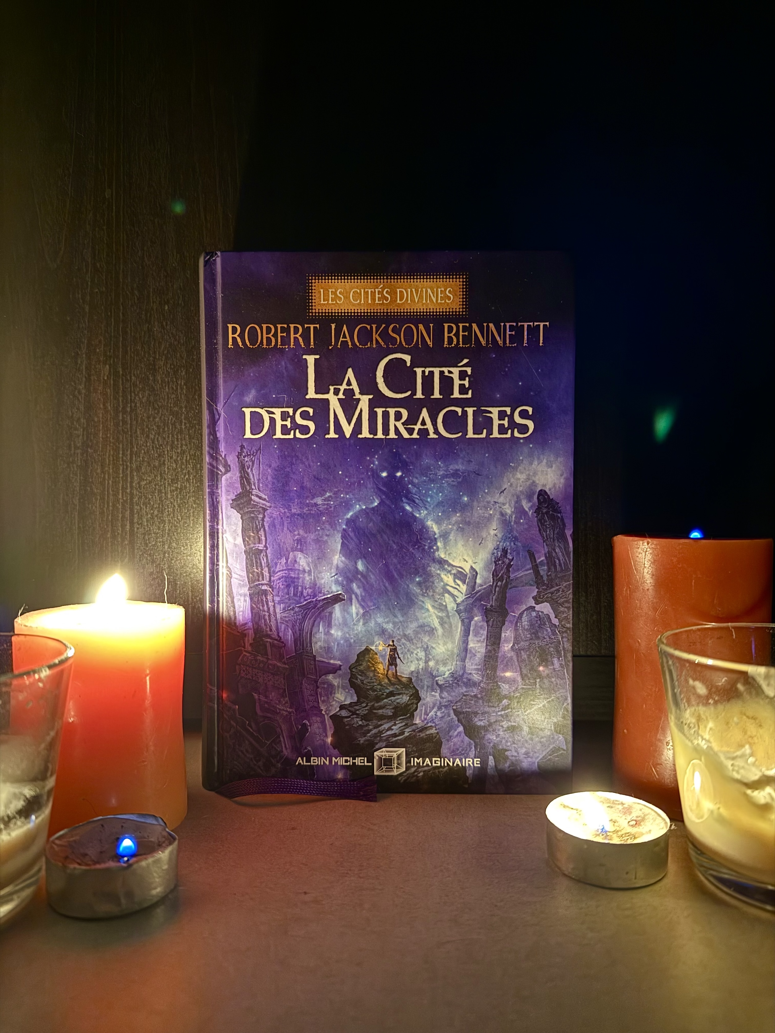 Livre posé dans la pénombre entouré de bougies allumées qui l'éclairent. La couverture de La cité des miracles montre une silhouette sur un promontoire en ruines, lanterne et lame à la main. Au coeur des ruines, une ombre gigantesque aux yeux brillants semble avancer vers la silhouette.