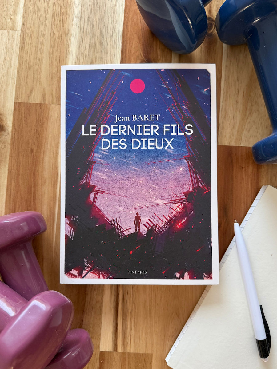 Livre Le dernier fils des Dieux posé aux cotés d'un carnet et bic et d'haltères. La couverture montre une silhouette au milieu de ruines en métal.