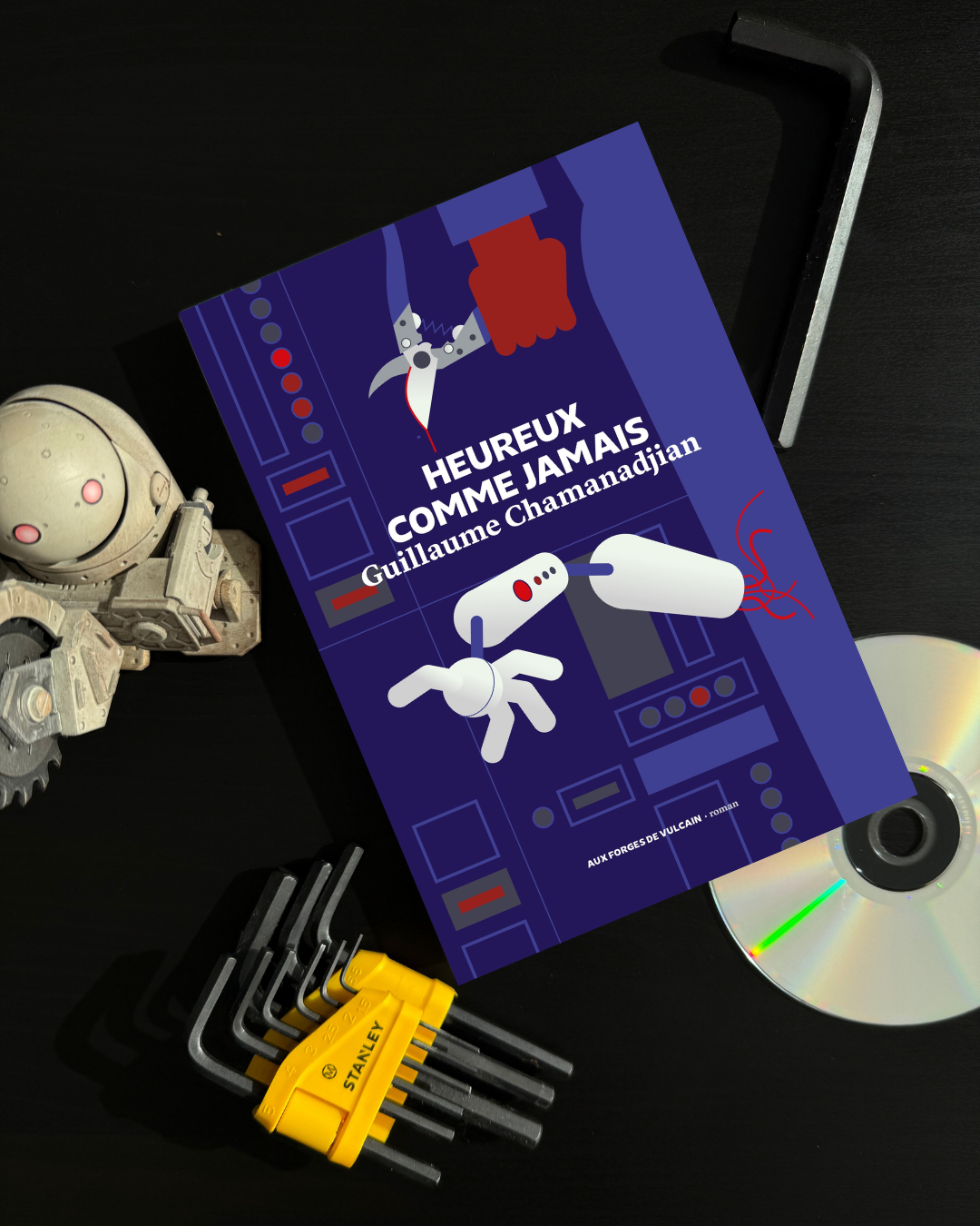 Livre posé aux côtés d'une figurine de robot tueur, d'un CD et de clefs Allen. La couverture d'Heureux comme jamais montre un corps en bleu de travail, pince coupante en main. Un bras robotique coupé chute à côté.