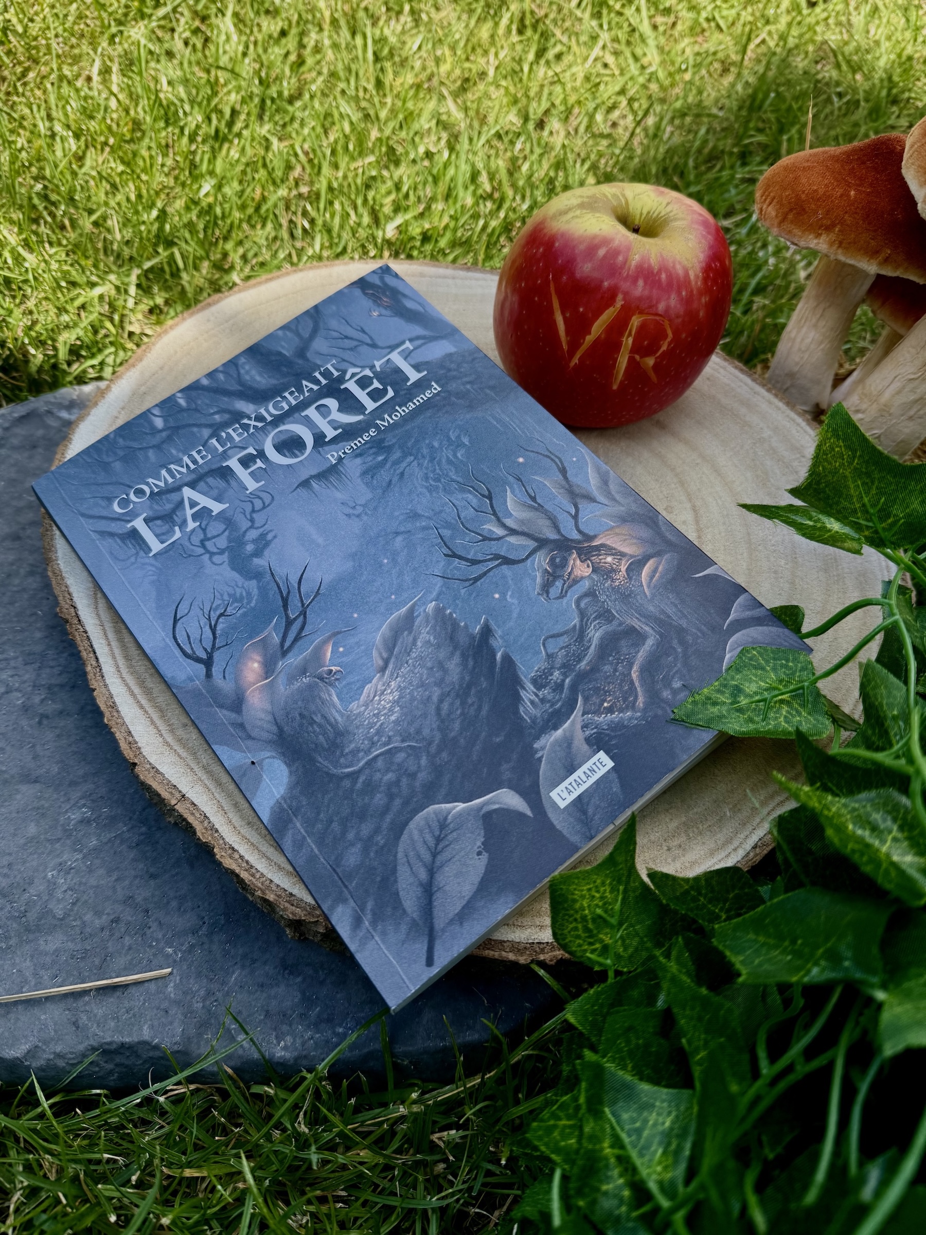 Livre posé dans l'herbe sur une souche d'arbre et une pierre avec du lierre et un champignon visibles. A ses côtés une pomme dans laquelle est gravée VR. La couverture de Comme l’exigeait la forêt montre des animaux à moitié squelettiques dont il se dégage de petites lueurs, dans bois sombres et inquiétants. 