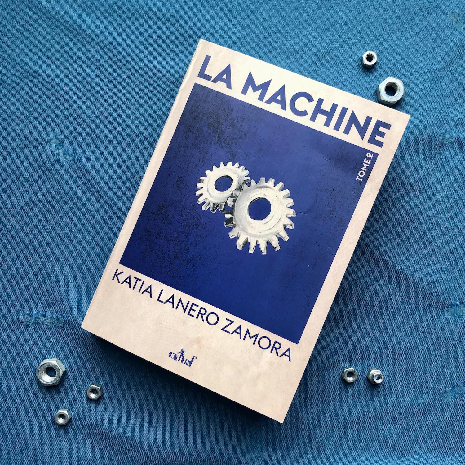 Le livre La machine, tome 2 de Katia Lanero Zamora chez ActuSf posé sur un tissu bleu sur lequel on voit des boulons. La couverture du livre montre un encadré bleu nuit sur fond beige. Dans ce cadre, deux rouages beiges sont illustrés au centre.