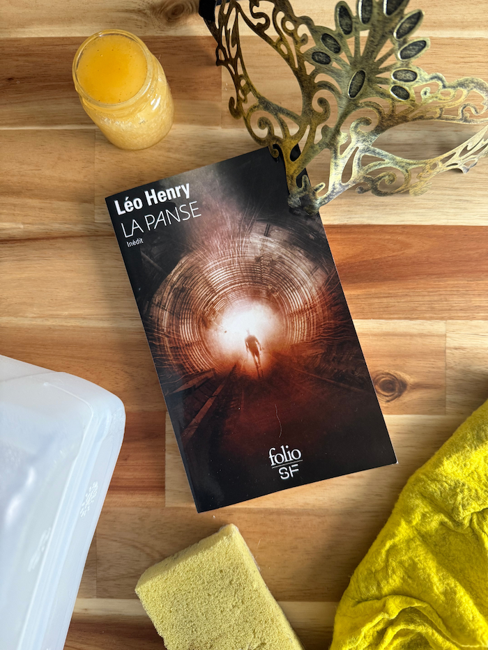 Livre La Panse avec un masque vénitien, un pot d’une substance jaune/dorée, et des ustensiles de ménage et un bidon de produit ménager