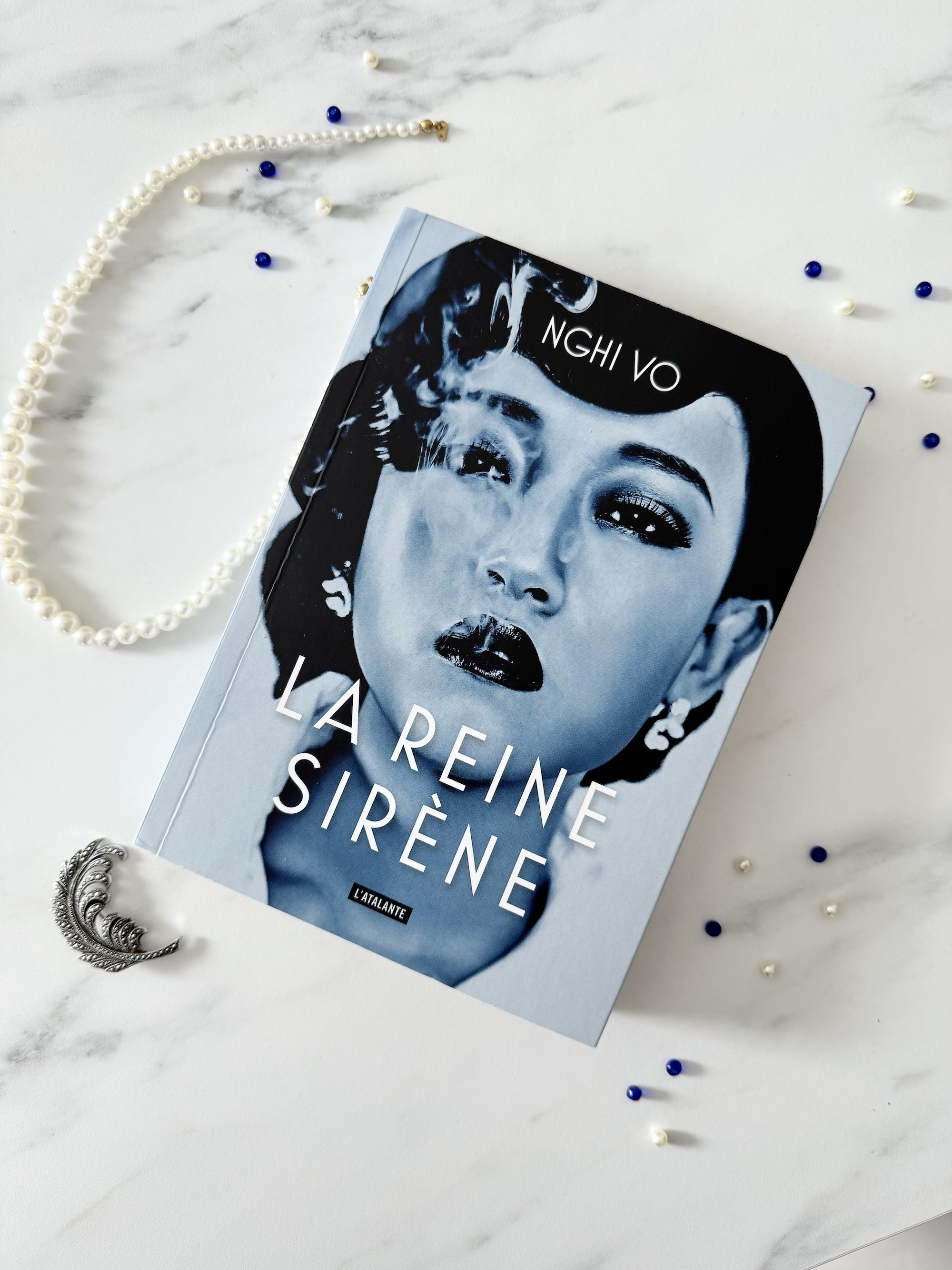  Livre posé à côté des perles bleus et blanches, d'une broche et d'un collier de perles. La couverture de La reine sirène montre un portrait de femme asiatique dans un ton bleuté/noir et blanc, maquillée, chignon et boucle d’oreilles, expulsant de la fumée de sa bouche entrouverte.