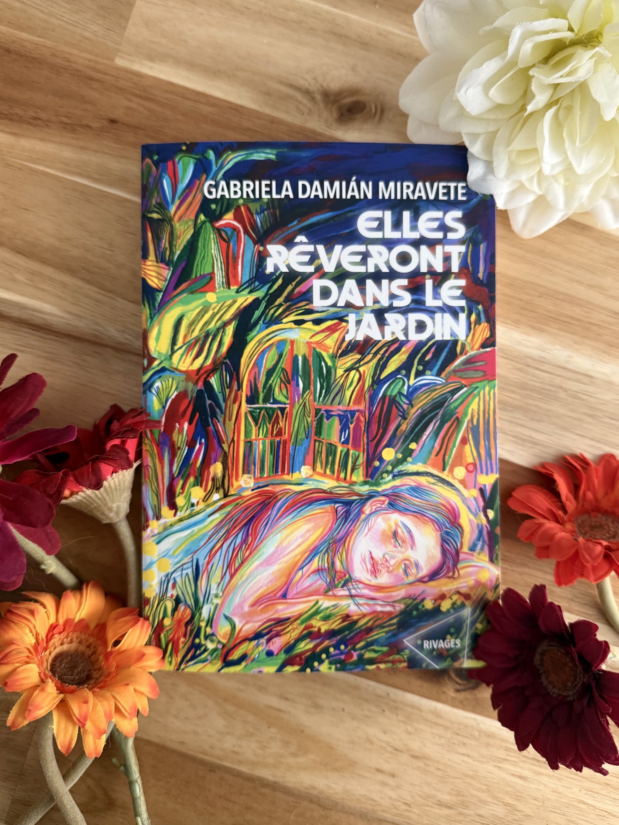 Livre posé au milieu de fleurs colorées. La couverture de Elles rêveront dans la jardin montre, dans des tons très vifs et colorés, une jeune femme endormie au milieu de plantes. En arrière-plan une fenêtre.