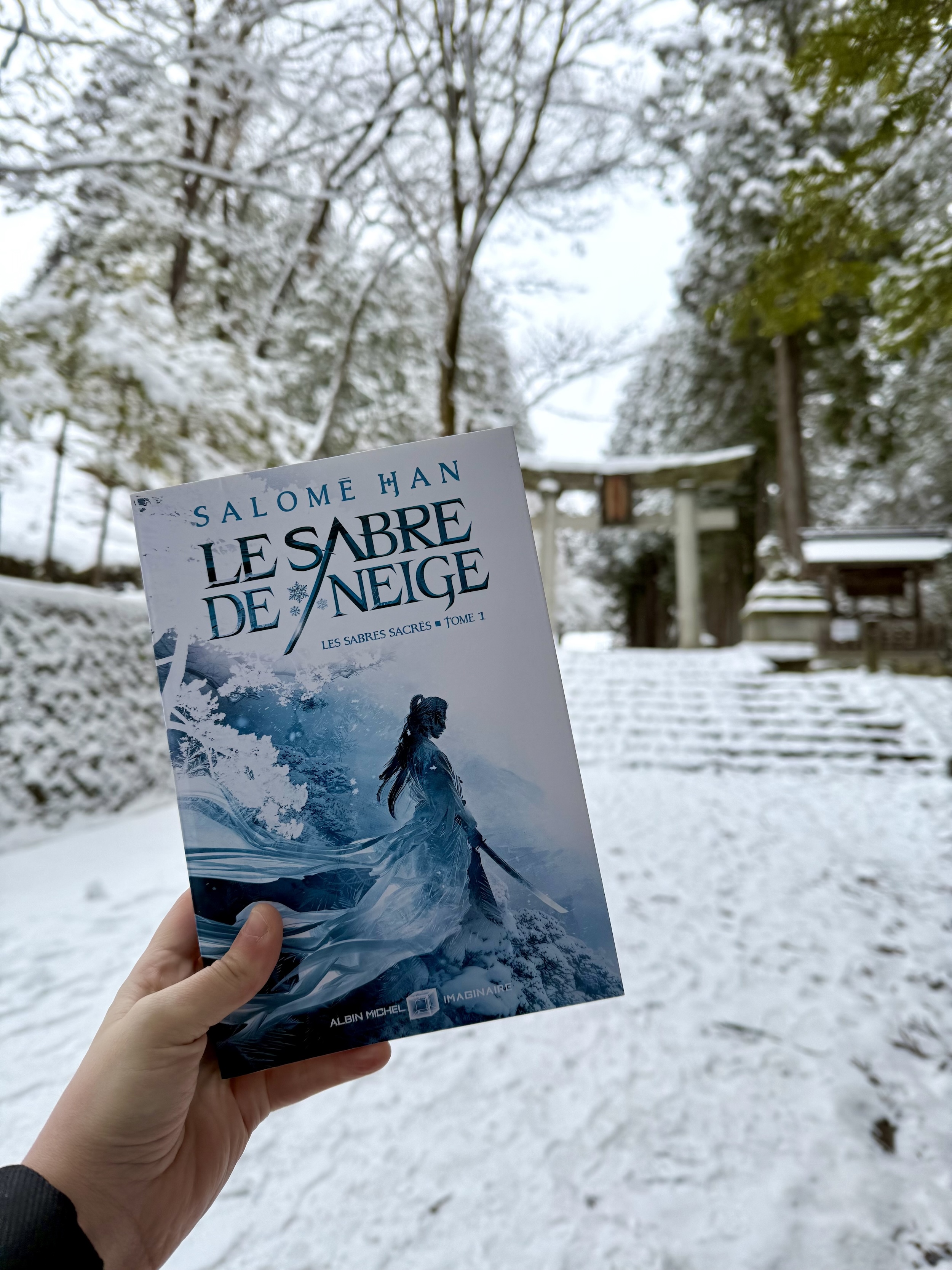 Livre pris en photo devant un torii de sanctuaire au Japon sous la neige (à Takayama si ça vous intéresse). La couverture du Sabre de neige montre un paysage enneigé avec un personnage, sabre en main, dont les tissus du vêtement couleur de glace s’envolent.